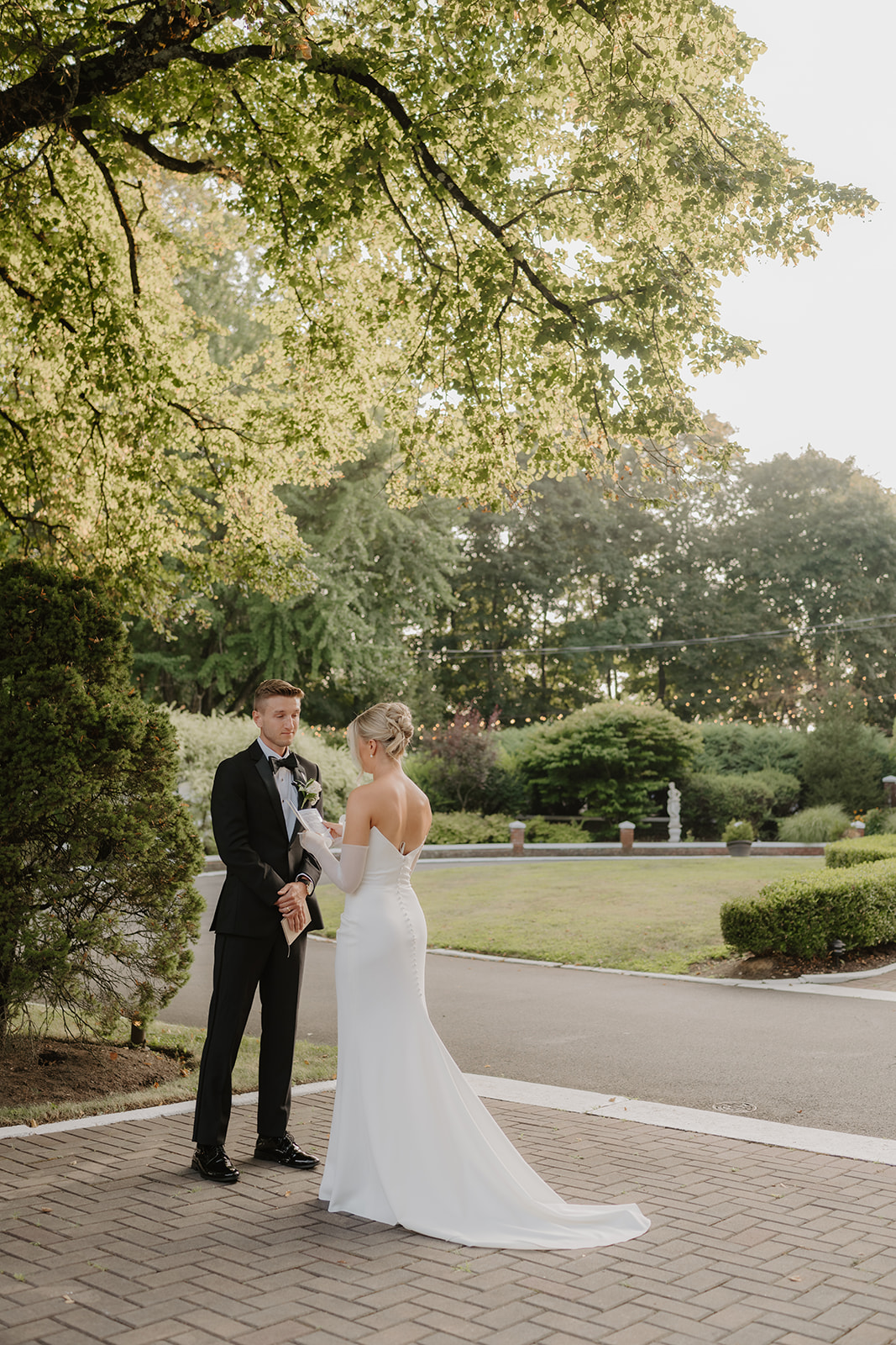 Newlyweds stand under a large tree on a brick path, reading vows to each other in a private ceremony.